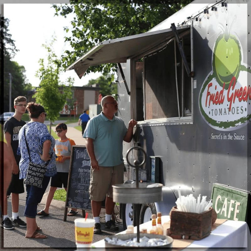 A family orders food from a "Fried Green Tomatoes" food truck, with a green "CAFE" sign and a table of condiments in the foreground, and trees and buildings in the background.