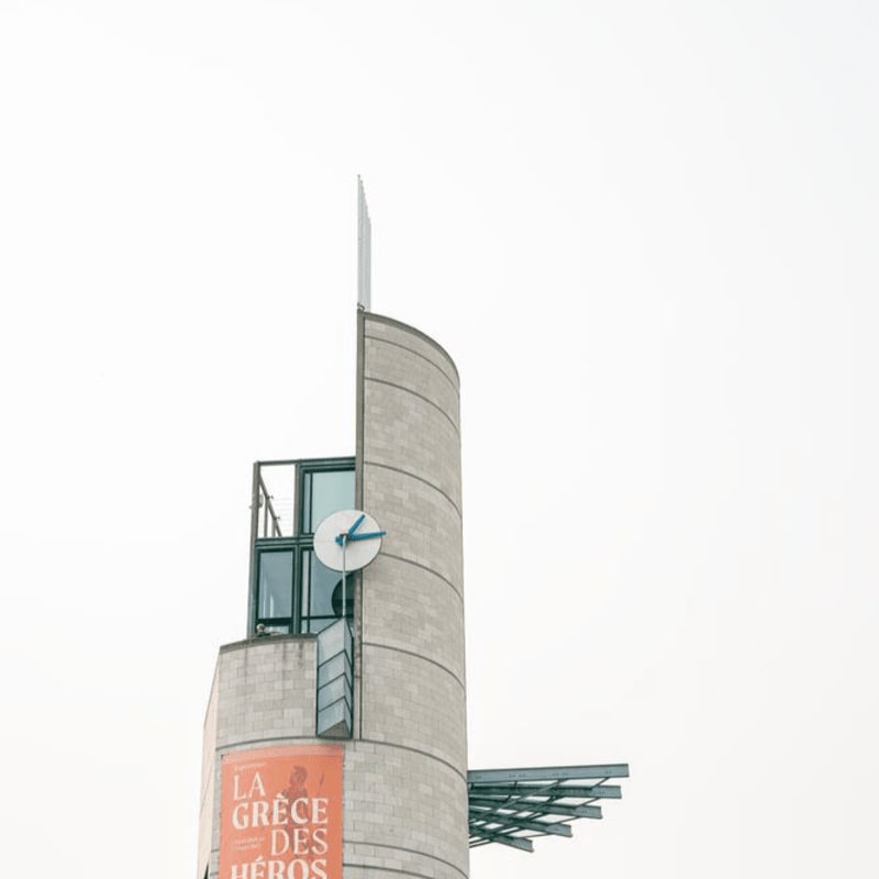 A cylindrical concrete tower with a clock face and an orange banner is set against a bright, overcast sky.