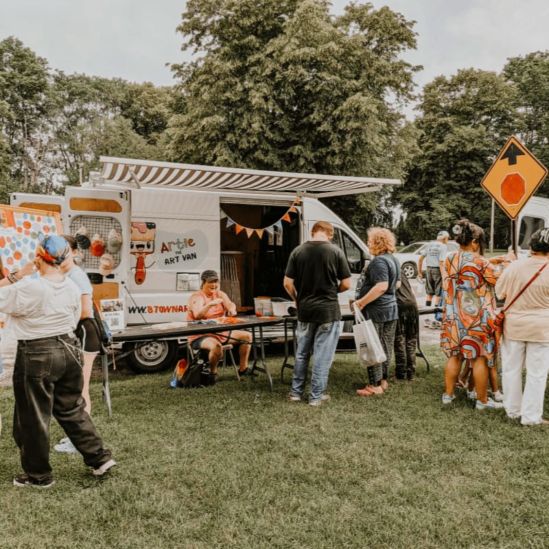 People gather around an art van with a striped awning in a grassy park, with trees and a road sign visible in the background.