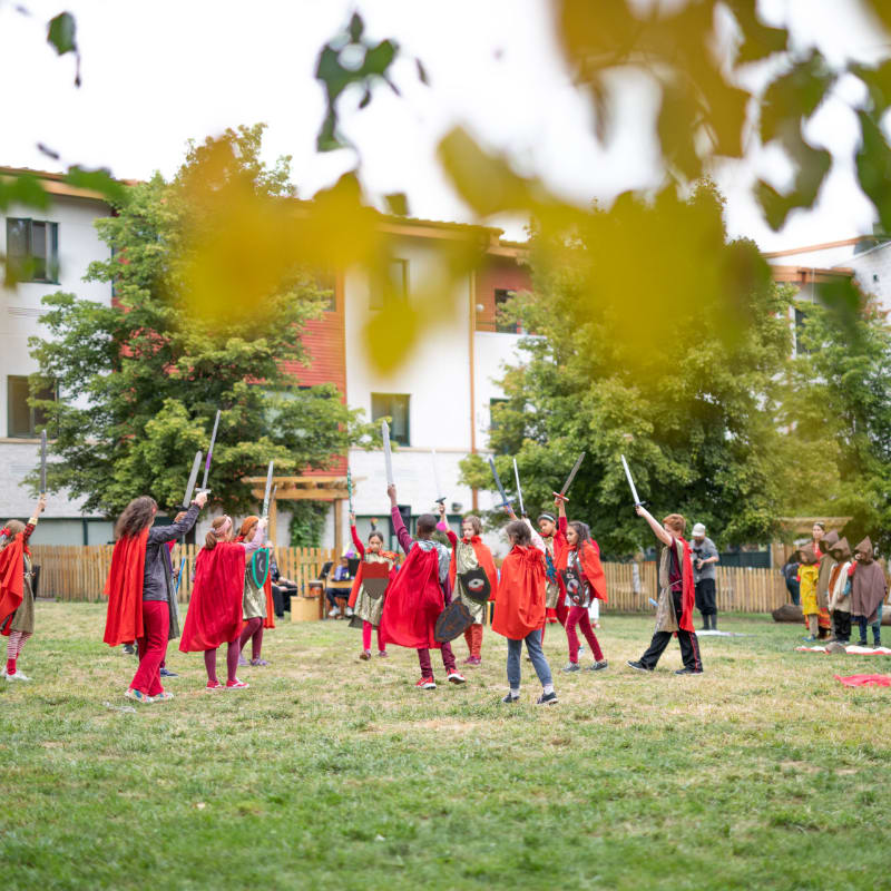 Children in red capes and holding swords engage in a playful sword fight on a grassy field, with a building and trees in the blurred background.