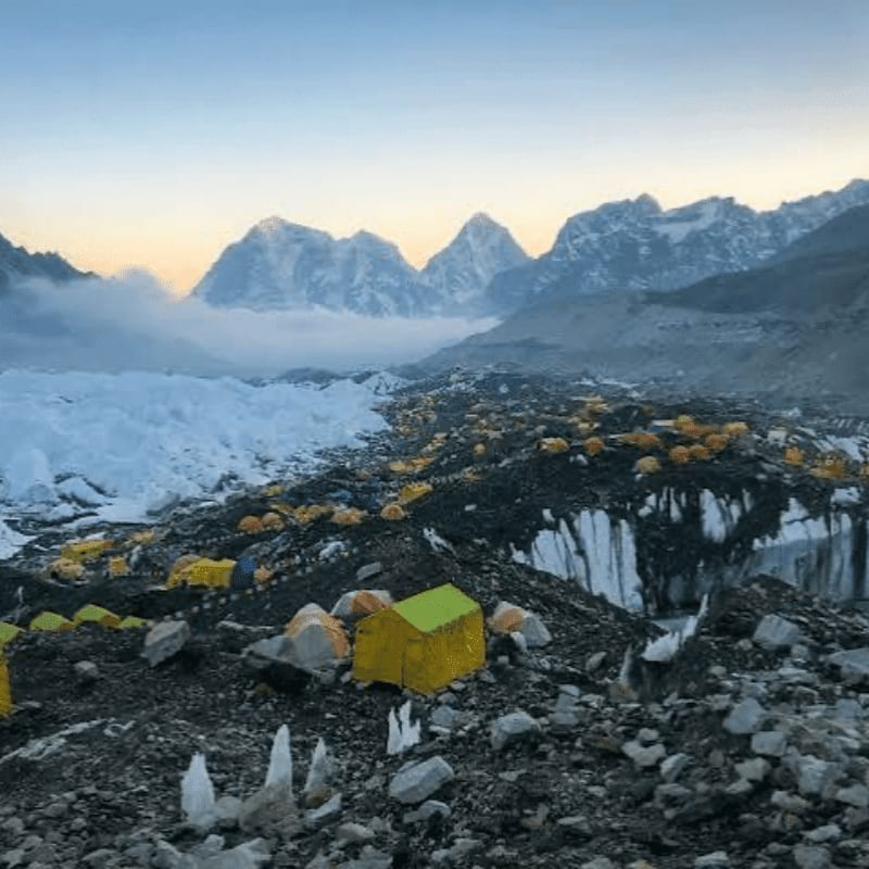 A vast tent city of yellow and blue shelters is spread across a rocky, icy foreground, with a majestic, snow-capped mountain range shrouded in mist in the background.
