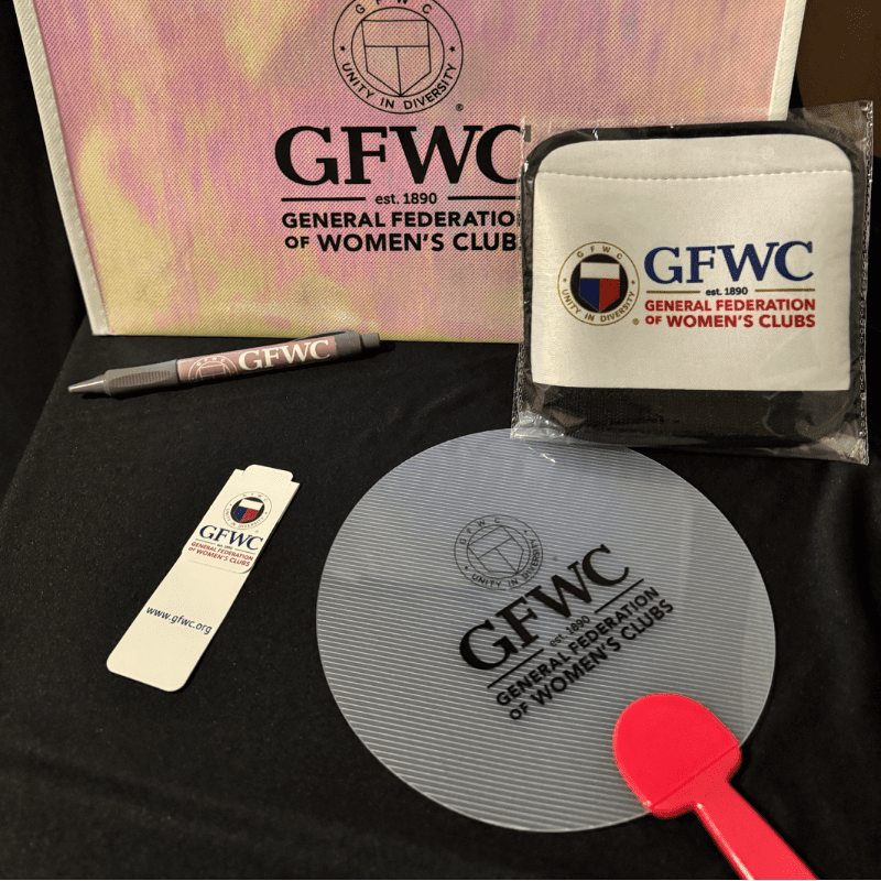 A pink tote bag and a pen are in the background, while a white pouch, a bookmark, a round fan, and a pink paddle are in the foreground, all featuring the GFWC logo.