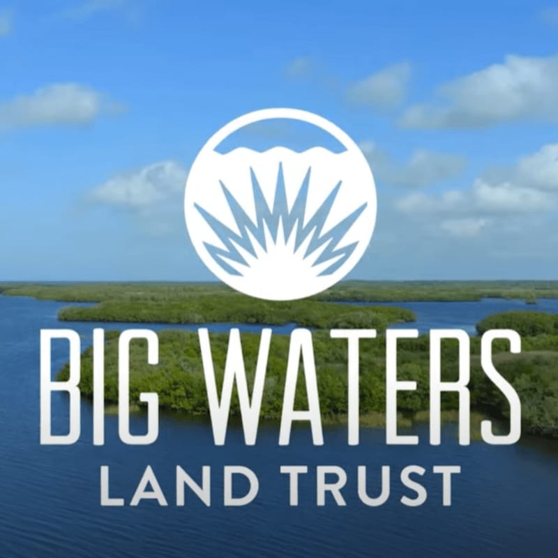 A circular logo with stylized water and sun rays hovers above the words "BIG WATERS LAND TRUST" which are superimposed on a tranquil blue body of water with lush green islands and a cloudy sky in the background.