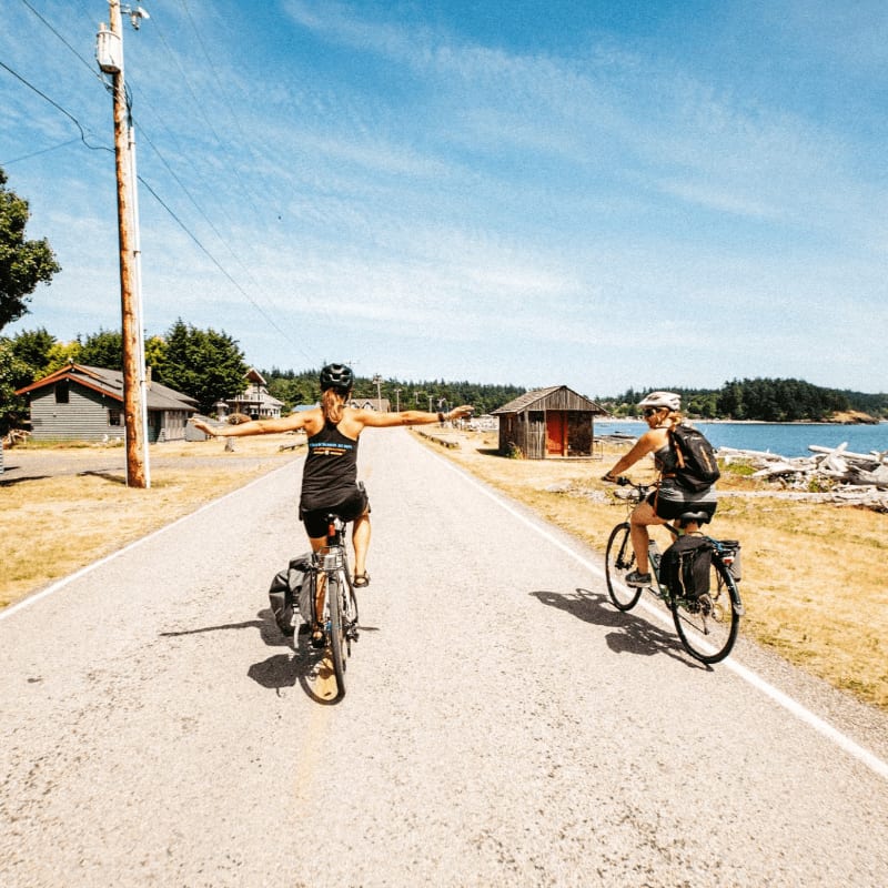 Two cyclists ride down a paved road with small buildings and trees on either side, leading to a body of water under a clear blue sky.