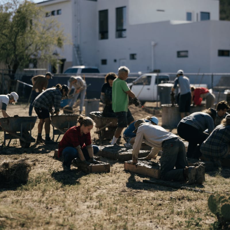 In the foreground, people are laying bricks on the ground, while in the background, a white building stands under a clear sky.