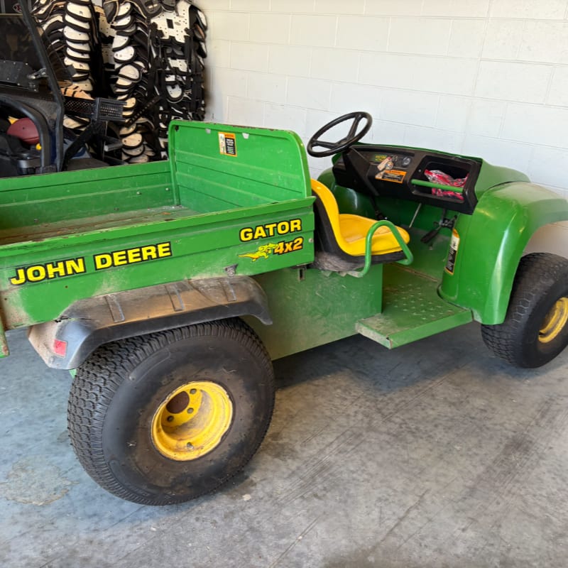 A green John Deere Gator 4x2 utility vehicle with yellow seats sits in a garage with stacked items in the background.