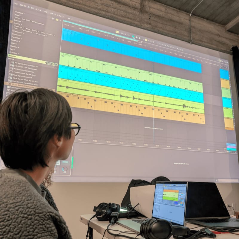 A person wearing glasses and a fuzzy jacket looks at a large projection screen displaying a music production software interface, with a laptop and headphones on a table in the foreground.