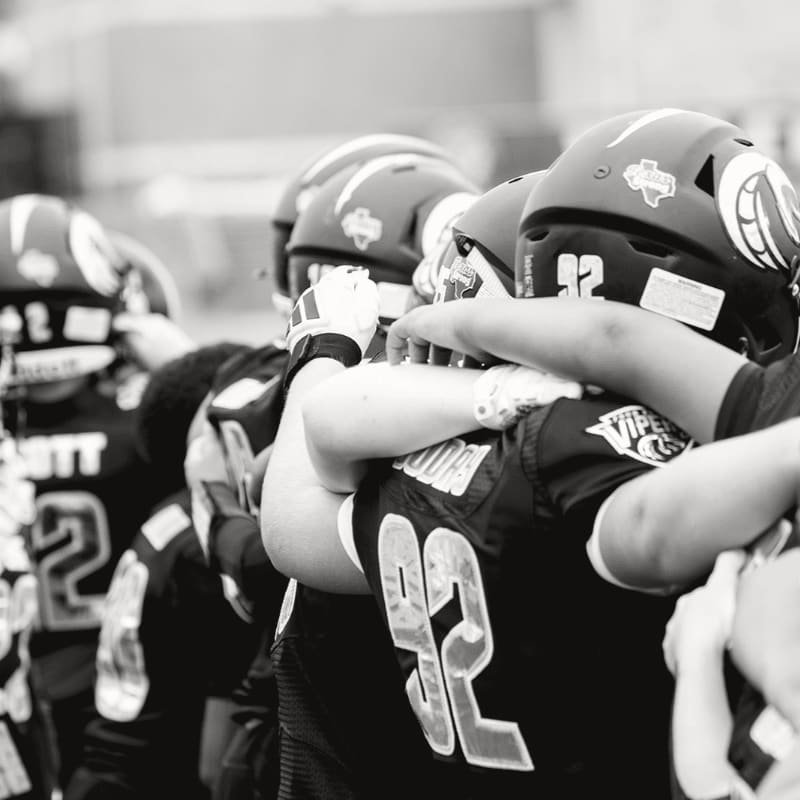 A line of American football players in uniform huddle together with their arms around each other's shoulders, their helmets and jerseys visible against a blurred outdoor background.