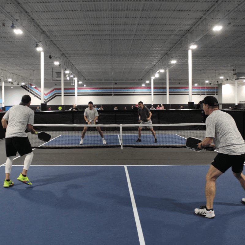 Four men play pickleball on a blue court with a white net, while spectators watch from the background of the indoor facility.