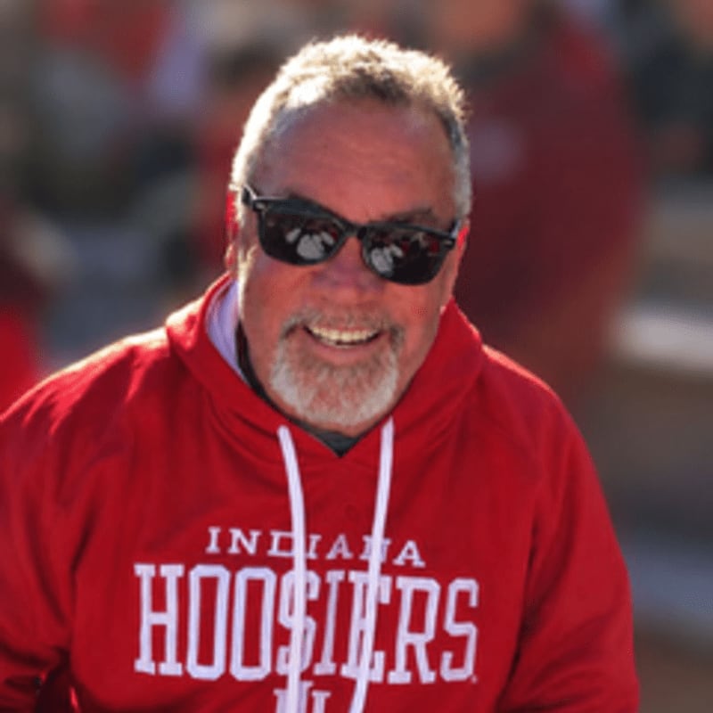 A smiling man in a red "Indiana Hoosiers" hoodie and sunglasses is in the foreground, with a blurred crowd in the background.