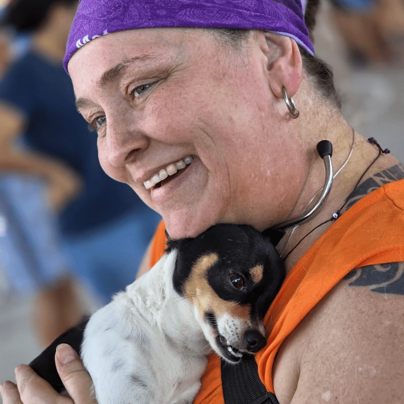 A smiling woman with a purple bandana holds a small dog close, with a blurred background of people.