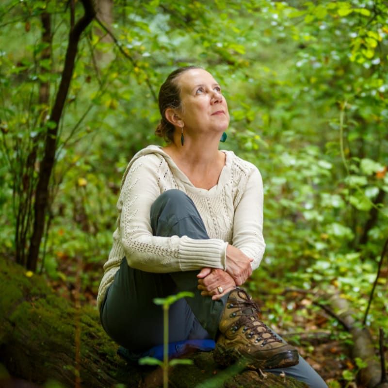 A woman in a cream-colored hoodie and hiking boots sits on a fallen log in a lush, green forest, looking upwards.