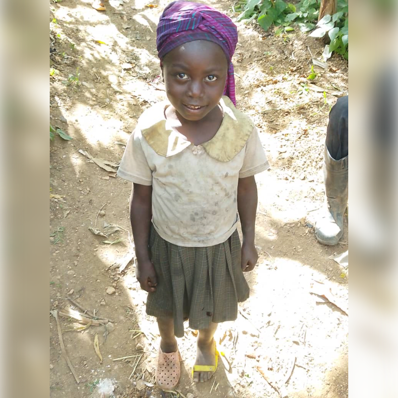 A young girl with a purple headscarf and a dirty shirt stands on a dirt path, with greenery and a person's legs in boots visible in the background.