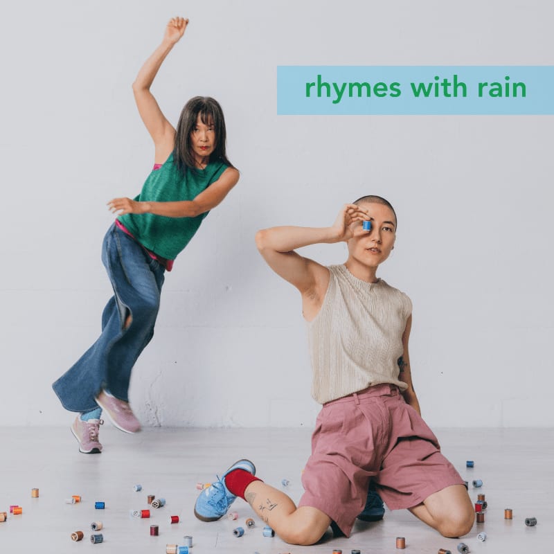 Two women dance amidst scattered colorful blocks on a white floor, with the text "rhymes with rain" in a blue box in the background.