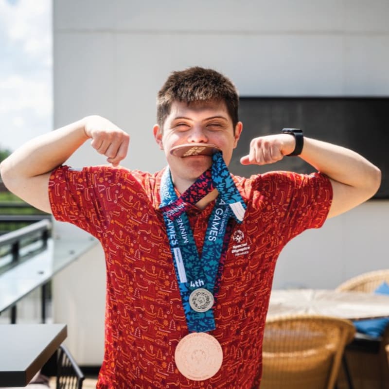 A young man in a red shirt flexes his biceps while holding medals in his mouth, with a blurred outdoor patio in the background.