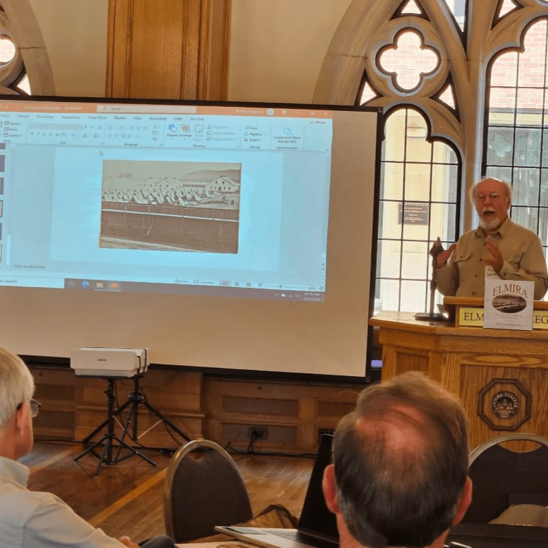 A presenter gestures while speaking at a podium in front of a projector screen displaying an old photograph, with attendees seated in the foreground and arched windows in the background.