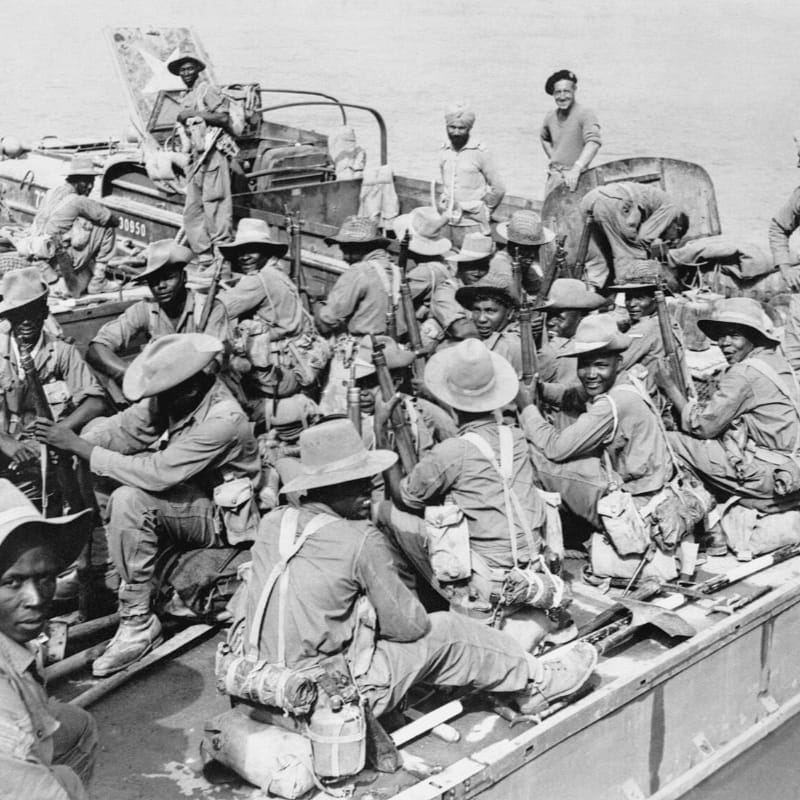 Soldiers in hats and uniforms sit packed together on a boat, with a calm body of water visible in the background.