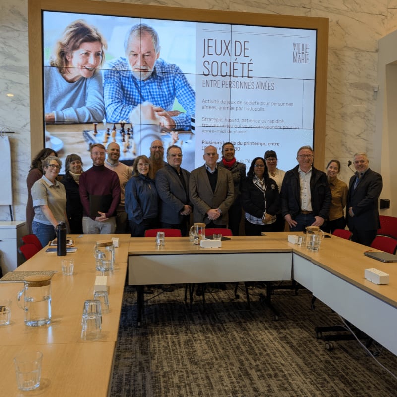 A group of people stand in front of a large screen displaying "JEUX DE SOCIÉTÉ ENTRE PERSONNES AINÉES" while a U-shaped conference table with glasses and pitchers sits in the foreground.