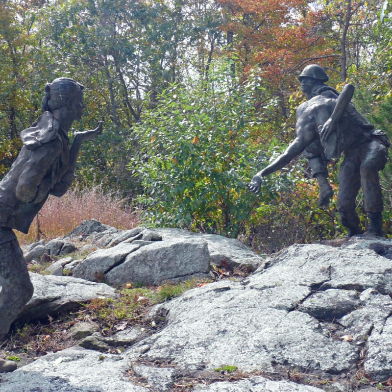Two bronze statues of a woman blowing a kiss and a soldier carrying a comrade stand on a rocky outcrop with autumn foliage in the background.