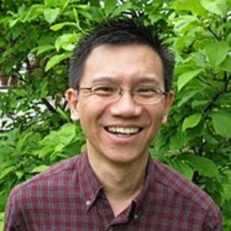 A smiling man in a plaid shirt is in the foreground, with lush green foliage in the background.