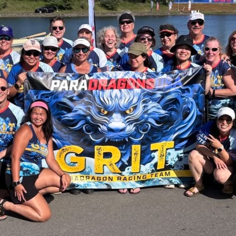 A group of smiling people in matching shirts pose in front of a banner featuring a dragon and the word "GRIT" with a body of water and trees in the background.