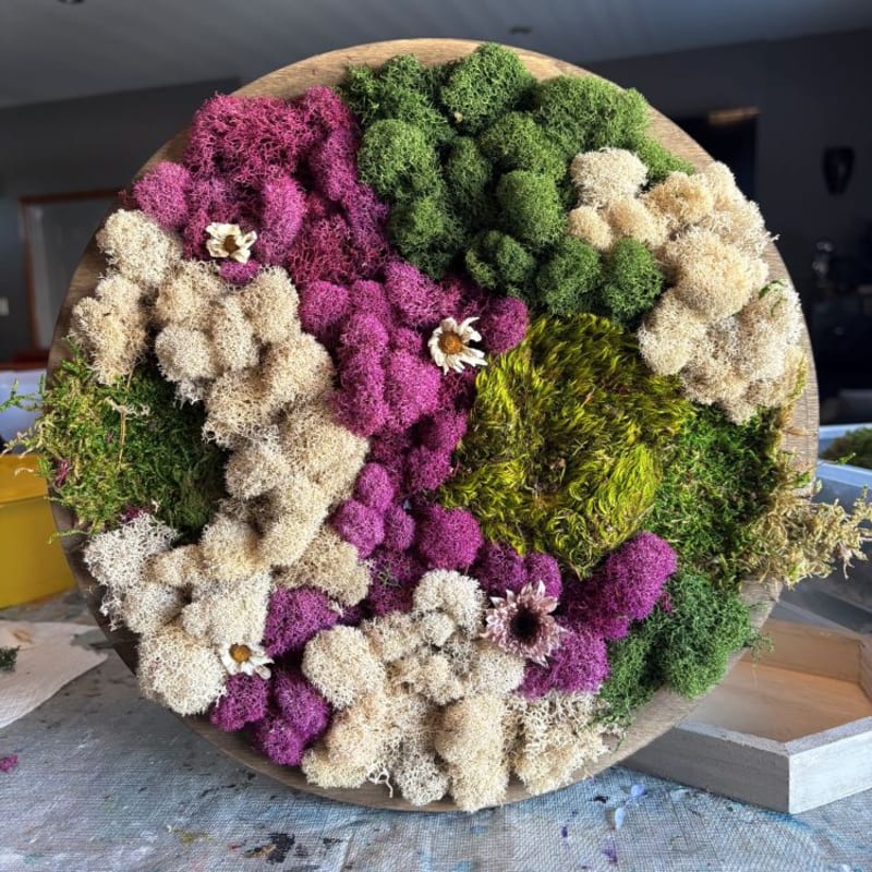 A circular arrangement of colorful preserved mosses and dried flowers sits on a table, with a blurred background of crafting supplies.