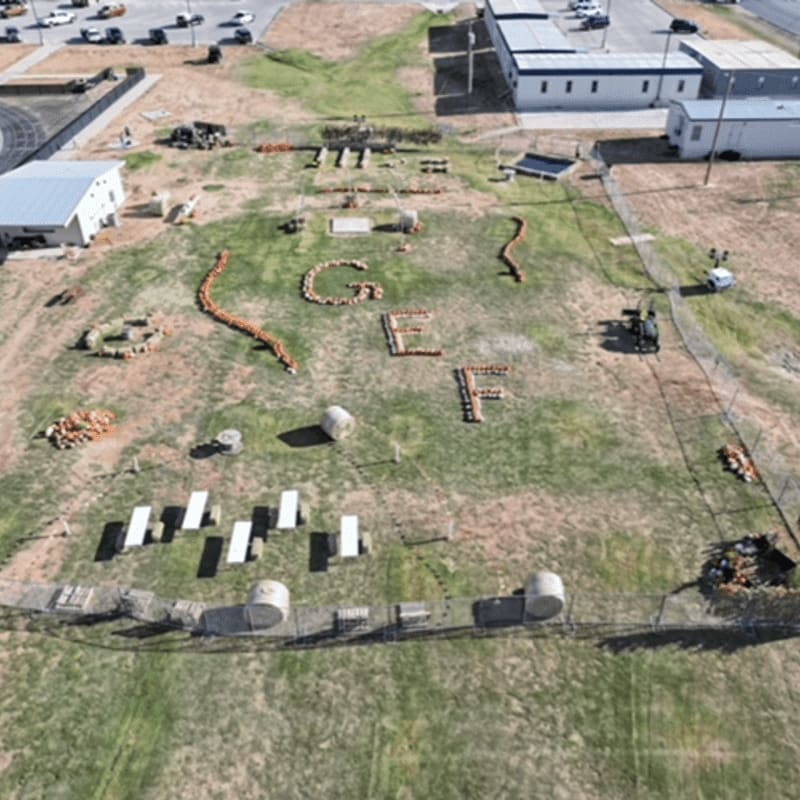 In the foreground, a grassy field is arranged with various objects and formations, including what appears to be letters spelled out with rocks and logs, while the background shows buildings and a parking lot.