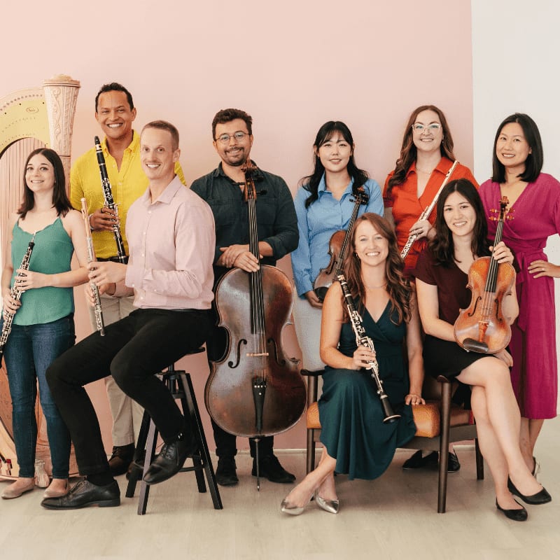 A diverse group of musicians, some holding instruments like violins and cellos, pose in front of a pale pink wall.
