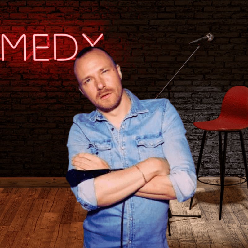 A man in a denim shirt stands with his arms crossed in front of a brick wall with a neon "COMEDY" sign and a microphone, while a red chair sits to the right.