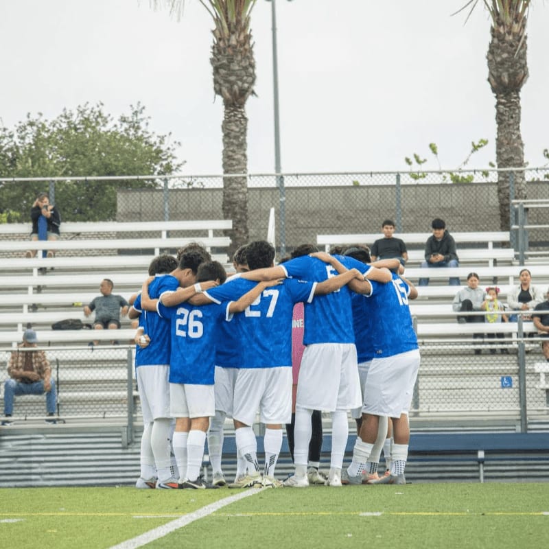 A soccer team in blue jerseys huddles together on a green field in front of bleachers filled with spectators and palm trees.