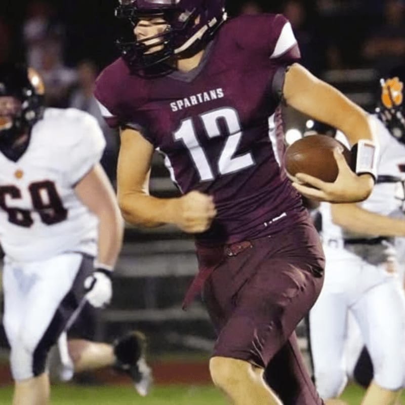 A football player in a maroon uniform with the number 12 on it runs with the ball in the foreground, while other players in white and orange uniforms are blurred in the background.