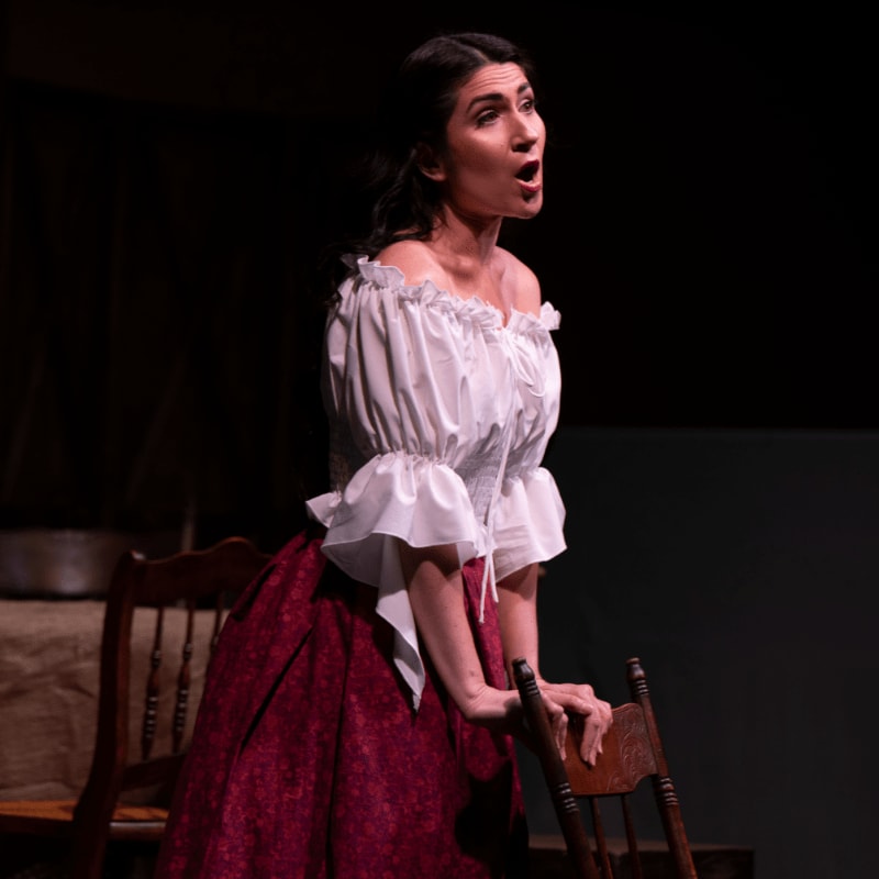 A woman in a white off-the-shoulder top and a burgundy skirt sings on a dark stage with wooden chairs in the foreground and a dimly lit backdrop.