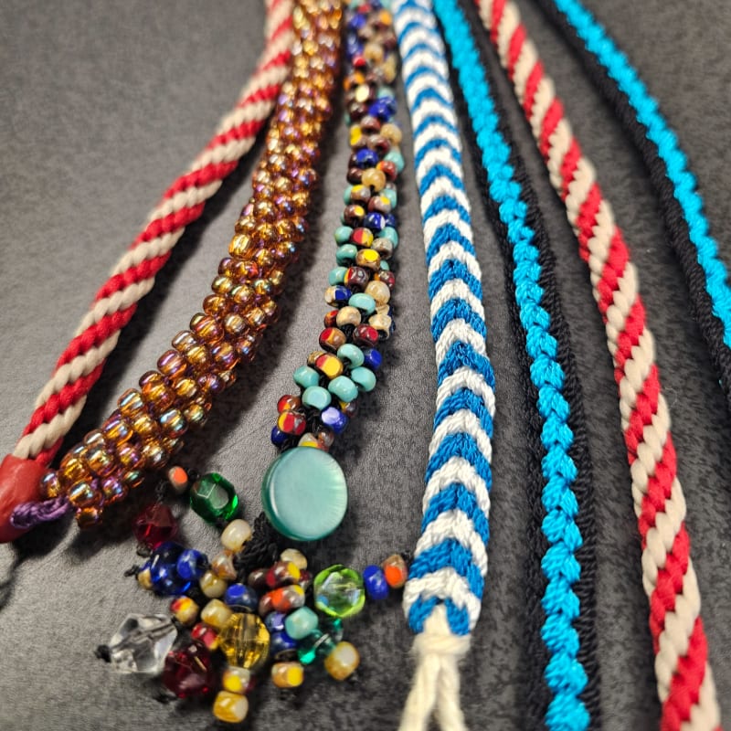 Several colorful, braided and beaded cords lie on a dark gray surface, with a cluster of beads and a turquoise pendant in the foreground.