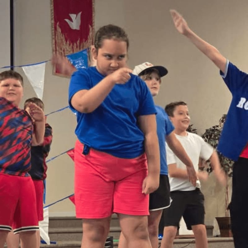 A group of children in casual attire are performing on a stage with red, white, and blue decorations in the background.