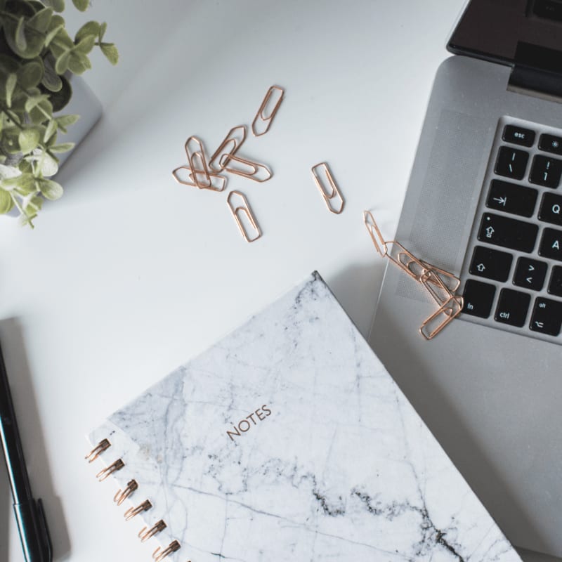 A marble-patterned notebook and scattered rose gold paperclips lie on a white desk next to a laptop and a potted plant.