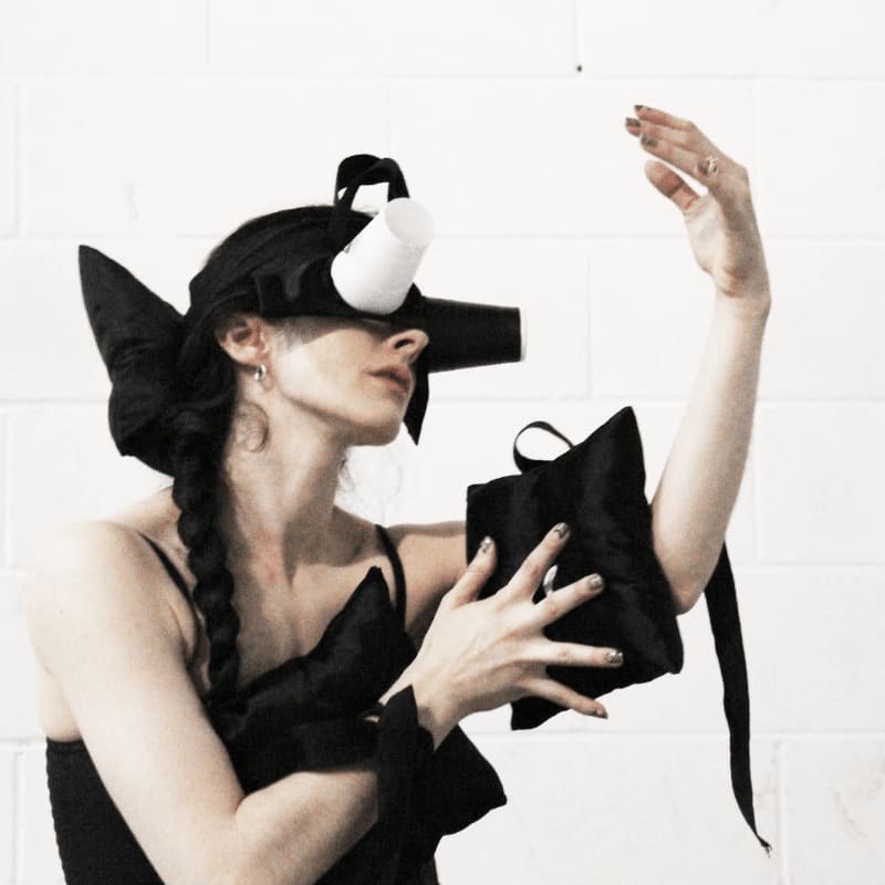 A person in a black avant-garde outfit and a unique headpiece with a white cup attached stands against a white brick wall.