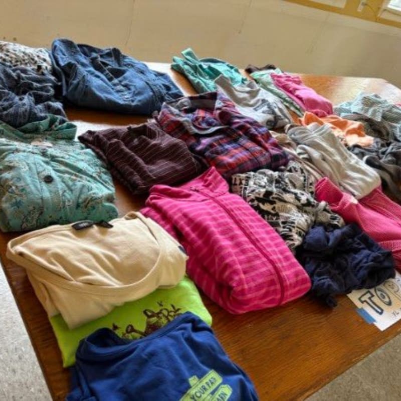 A collection of folded and piled clothing, including shirts and sweaters, is spread across a wooden table with a window visible in the background.