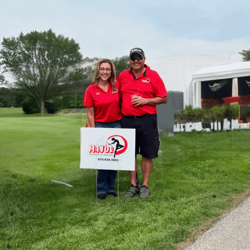 A man and woman in red shirts stand in a grassy field holding a sign, with a white tent and trees in the background.