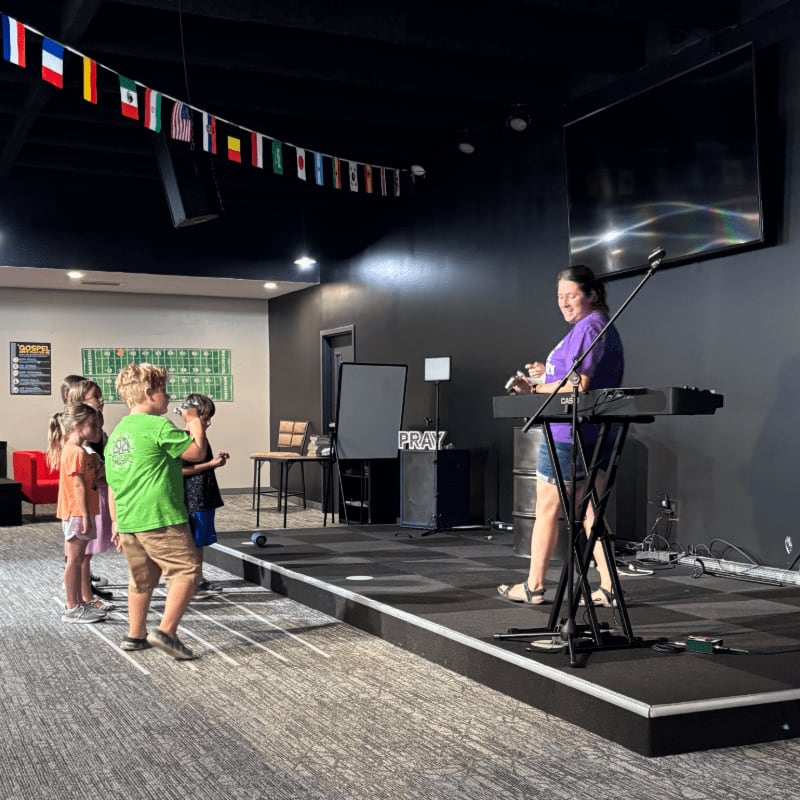 A group of children stand on a carpeted floor in front of a stage where a woman plays a keyboard, with flags strung across the dark ceiling in the background.