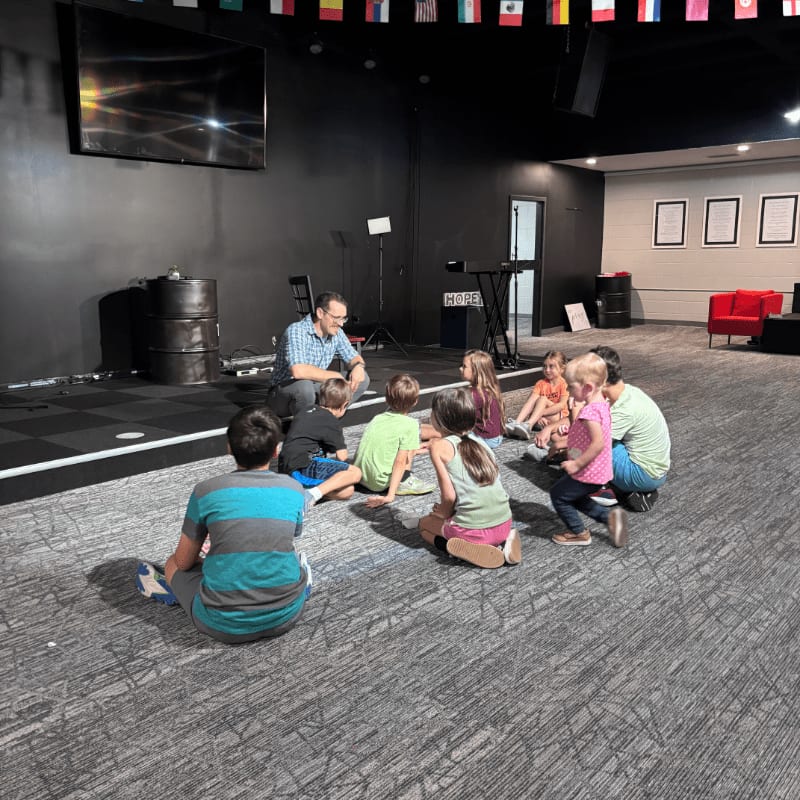 Children and adults sit in a circle on a patterned carpet in the foreground, while a stage with a large screen and flags hangs in the dark background.