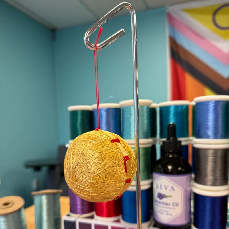 A golden yarn ball hangs from a metal stand in the foreground, with spools of colorful thread and a bottle of lavender oil in the background.
