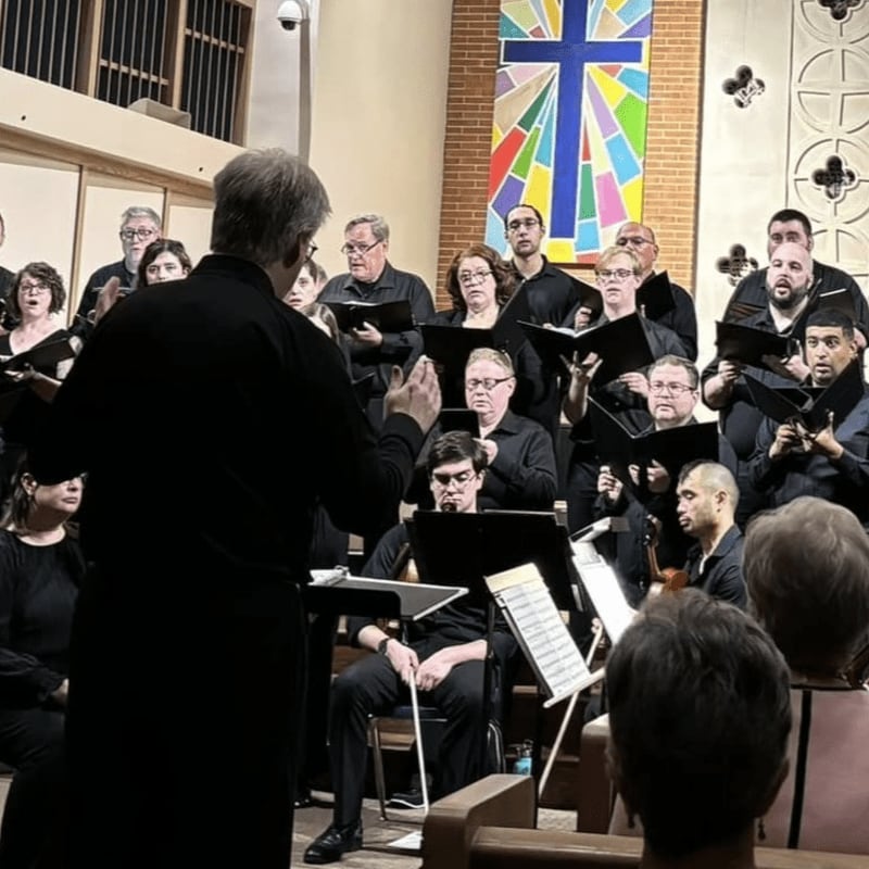 A choir in black robes sings in front of a colorful stained-glass window and wooden pews.