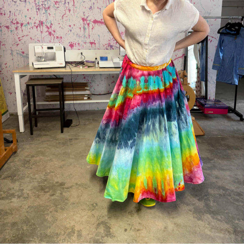 A person stands in a studio wearing a vibrant tie-dye skirt, with a sewing machine and art supplies visible in the background.