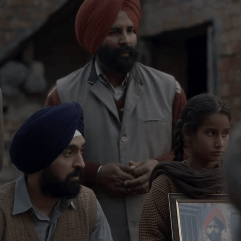 In the foreground, a young girl holds a framed photograph of a man in a turban, while two men in turbans stand behind her, their faces somber against a blurred, rustic background.