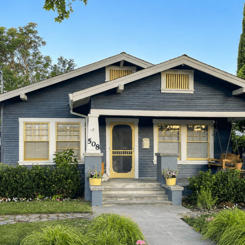 A charming blue bungalow with a yellow screen door and porch swing sits behind a manicured lawn and walkway, with lush green bushes framing the front of the house under a clear blue sky.
