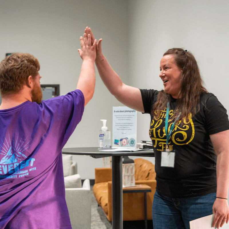 Two people high-five in the foreground, while a table with a sign and hand sanitizer sits in the blurred background.