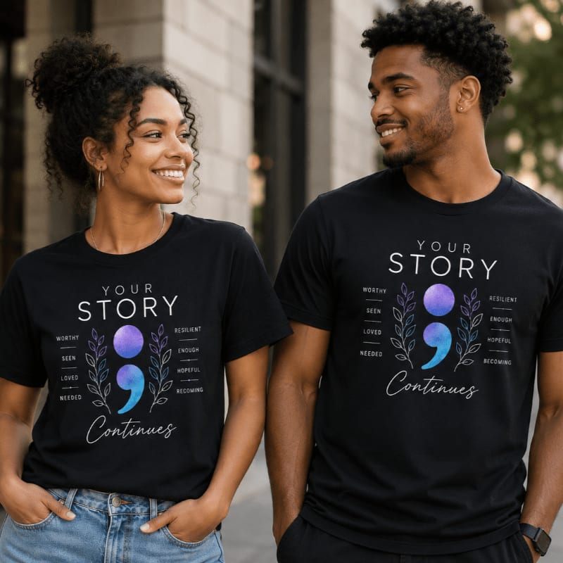A smiling Black couple stands close together, their black t-shirts featuring a semicolon design and empowering words, with a blurred urban background behind them.