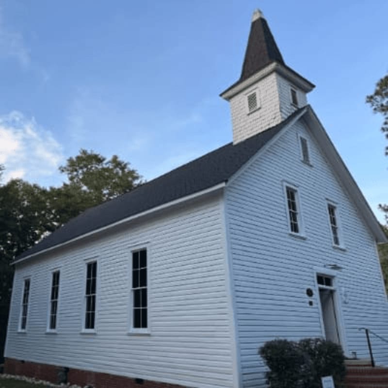 A white wooden church with a steeple stands against a clear blue sky, surrounded by lush green trees.