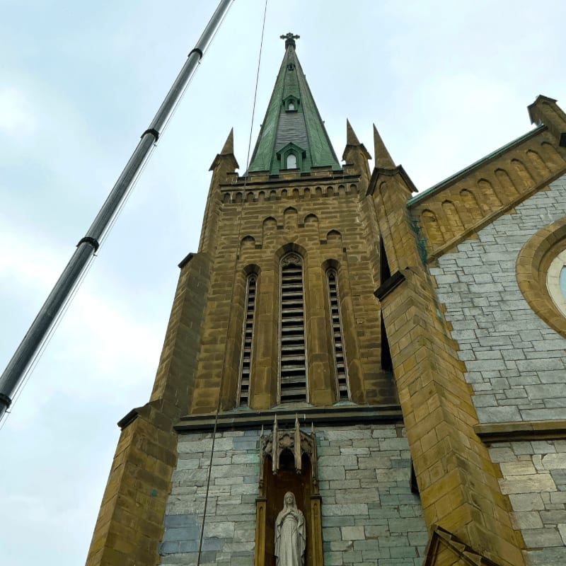 A crane boom extends from the left foreground towards the sky, framing a stone church with a tall, green-capped spire and a statue of a religious figure in a niche.