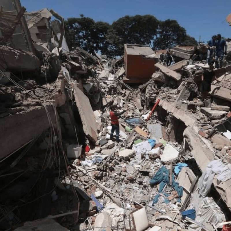 A lone figure stands amidst the rubble of a collapsed building, with trees visible in the clear blue sky behind.